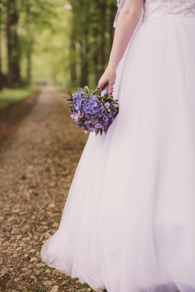 mariée sur un chemin de terre tient son bouquet violet à la main