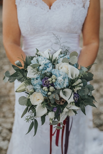 Bouquet de mariée champêtre avec nombreux feuillages et pommes de pins, tons blancs, bleus et violets.